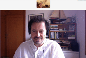 A screenshot of a video call featuring two people. The main participant, Blas Subiela, a man with dark curly hair and a beard, is wearing a white shirt and black headphones. He is seated in a room with a wooden bookcase behind him, which contains books, model sailboats, and various decorative items. The second participant, Pilar Orero, appears in a smaller window at the top of the screen and is a woman wearing glasses and a striped top, smiling at the camera.