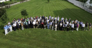 A large group of international participants stands on a green lawn, posing for a group photo during the 2nd UN Virtual Worlds Day in Turin, Italy. The photo includes men and women of diverse backgrounds, some wearing blue ITU shirts, with trees and a fence visible in the background. The ITU logo is in the top right corner.