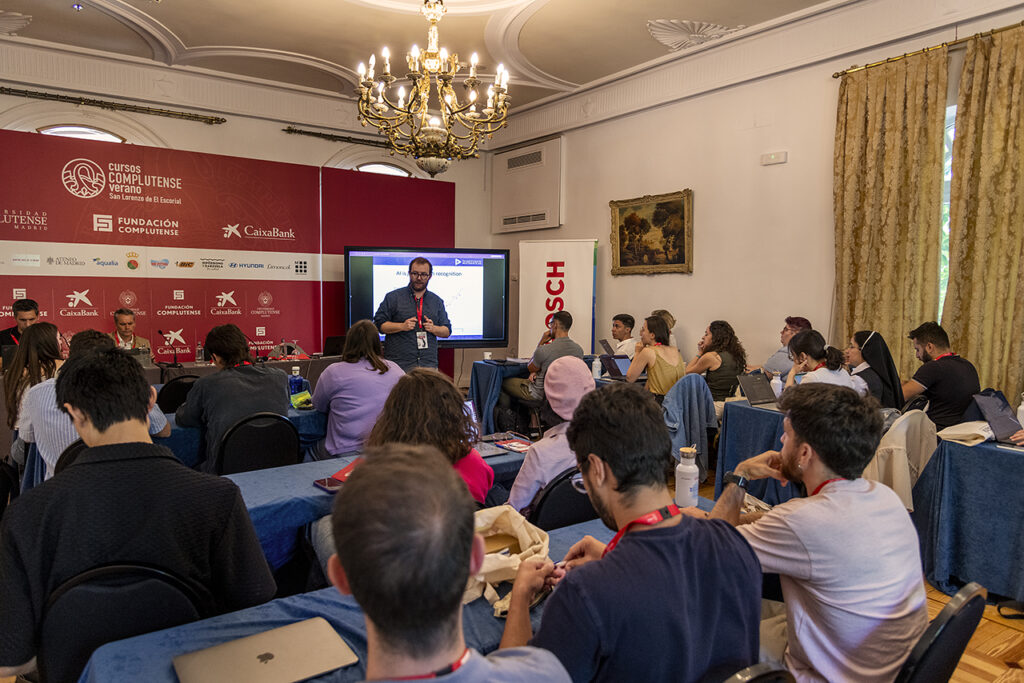 A classroom setting during a summer course at Universidad Complutense de Madrid, with a speaker presenting on AI in sensory recognition to a group of attentive students seated at tables with laptops; the room features elegant decor, a chandelier, and sponsor banners including Bosch.