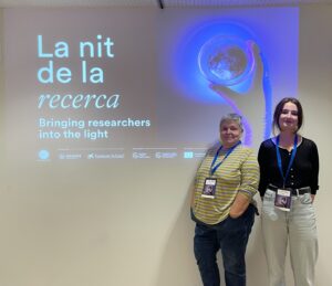 1- Two women standing side by side in front of a CosmoCaixa projection that reads “La nit de la recerca / Bringing researchers into the light.” On the left: Pilar Orero, wearing a yellow-and-green striped long-sleeve shirt and jeans; on the right: Sarah Anne McDonagh, wearing a black top and light-colored pants.