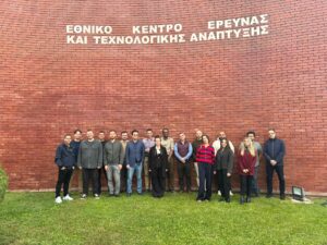 ALFIE project partners standing together outside the CERTH headquarters in Thessaloniki, Greece, in front of a red-brick wall with the Greek sign for the National Centre for Research and Technology Development.