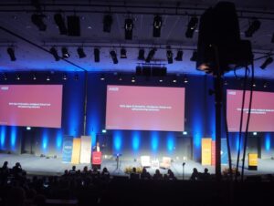 Alt text: “Wide-angle view of the AIS25 conference stage in a large auditorium. Three large projection screens display presentation slides with red backgrounds. A speaker stands at a podium center-left, two chairs and a small table are arranged stage-right. The audience silhouettes are visible in the foreground and blue stage lighting washes the back wall. Overhead stage lights and suspended equipment are visible near the ceiling.”