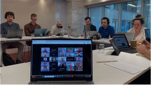 A hybird meeting setup for the ALFIE project, showing a laptop screen in the foreground displaying a video conference with multiple remote participants, while in-person attendees sit with their laptops in a conference room in the background.