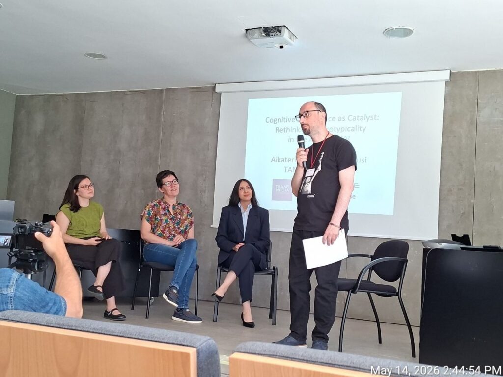 A snapshot from the TANC 2026 conference panel discussion. Co-developer Haris Shekeris stands speaking into a microphone, while panel discussants sit on stage next to a projector screen displaying Dr. Aikaterini Tsaousi's presentation on neurodiversity and AI.