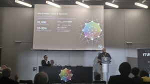 A speaker stands at a podium delivering a presentation at an ITU Kaleidoscope conference. Behind him, a large projection screen displays a slide titled "Why It Matters: Safety & Regulation." The slide contains statistics on drowsy driving (91,000 US crashes annually) and details about the EU Mandate (GSR 2019/2144), alongside a colorful, geometric network graphic. Another panelist sits at a desk to the left, and the backs of audience members are visible in the foreground.
