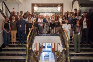 A large group of summer school attendees pose for a group photo on a grand marble staircase inside an ornate building lobby.