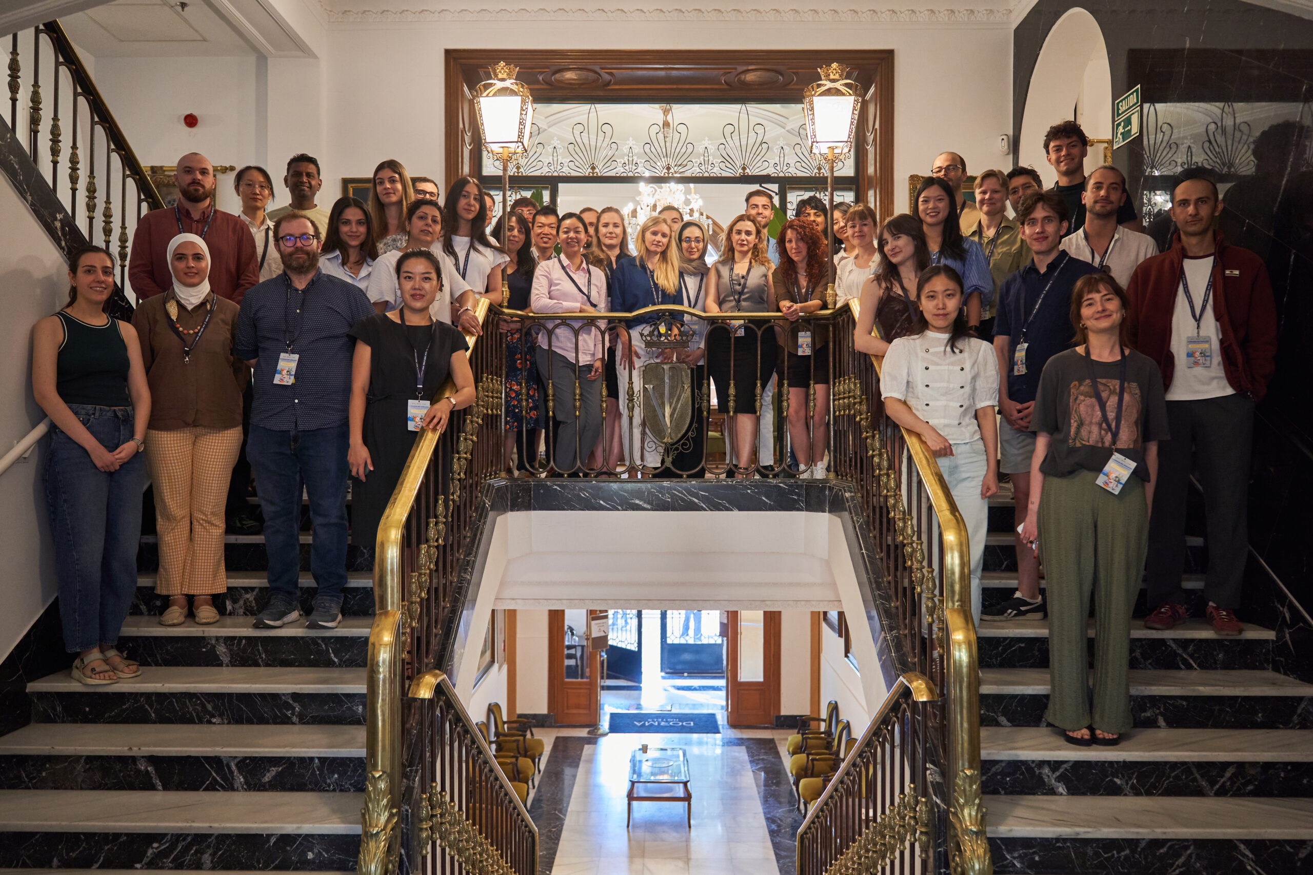 A large group of summer school attendees pose for a group photo on a grand marble staircase inside an ornate building lobby.