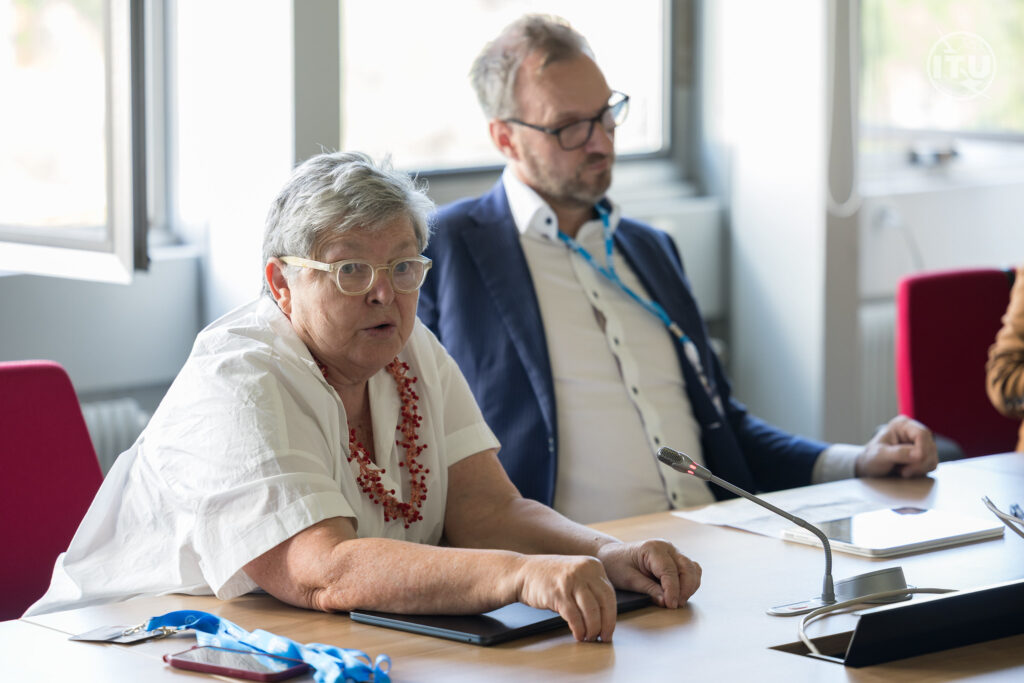 A close-up shot of Professor Pilar Orero speaking during a panel session at a conference table. She is wearing a white blouse and a red beaded necklace. Seated slightly behind her is a male colleague in a blue suit jacket and glasses, listening as she speaks. A tabletop microphone sits in front of them, and an ITU logo is visible in the top right corner.