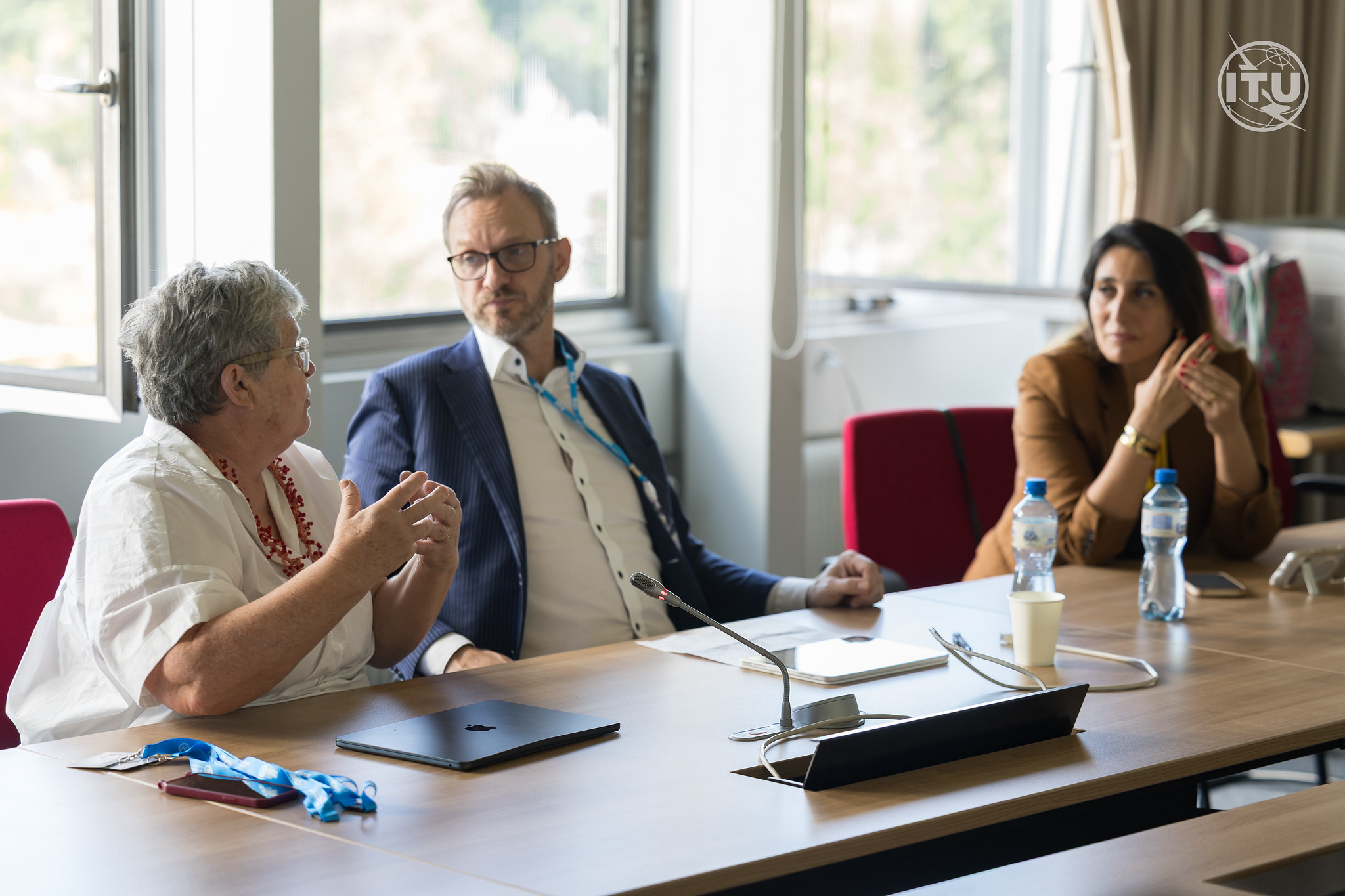 A side-profile view of Professor Pilar Orero gesturing with her hands as she speaks during a workshop panel. Seated next to her, a man in a blue suit jacket looks towards her attentively. Further down the table, a woman in a brown blazer listens with her hands clasped. The setting is a brightly lit conference room with large windows and an ITU logo in the upper right corner.